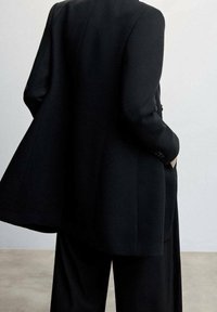 Zwarte op maat gemaakte blazer met een brede snit, gestructureerde stof en verlaagde schouders, gecombineerd met bijpassende wijde broekspijpen. Minimale accessoires.