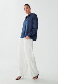 Camicia satinata blu con bottoni, maniche lunghe e colletto a punta, abbinata a pantaloni bianchi a gamba larga e tacchi neri con cinturini.