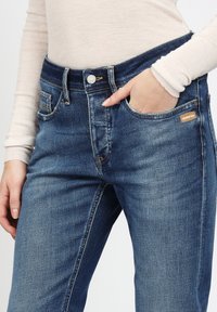 Dunkelblaue Denim-Jeans mit Knopfverschluss, Gürtelschlaufen, Seitentaschen und einem kleinen Markenschild an der rechten Vorderseite der Tasche.
