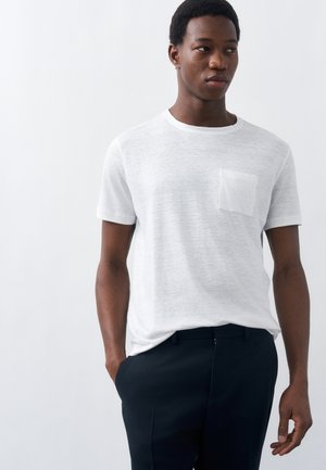 Νεαρός άνδρας με κοντά σγουρά μαλλιά, που φοράει λευκό T-shirt με τσέπη και μαύρο παντελόνι, στέκεται με το ένα χέρι στην τσέπη σε απλό φόντο.