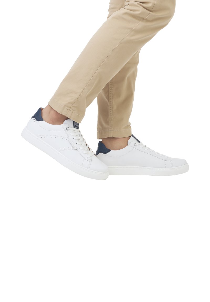 Sneakers bianchi con dettagli blu navy, pelle liscia, design perforato sui lati e suola piatta in gomma, indossati con pantaloni beige chiaro.