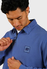 Pullover blu con zip in tessuto morbido, caratterizzato da un colletto alto e un'etichetta con logo di un elefante. Il cursore della zip è blu, abbinato al colore del capo.