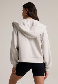 Sudadera gris claro con un corte holgado, gran capucha y dobladillo acanalado. Presenta mangas largas ligeramente fruncidas y una tela suave y lisa.