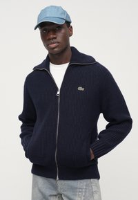 Tmavomodrý pletený sveter na zips s vysokým golierom, zdobený malým zeleným logom, doplnený svetlom modrou džínovou šiltovkou.