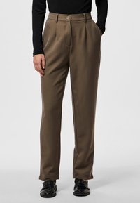 Braune maßgeschneiderte Hose mit hoher Taille, gefaltetem Frontbereich und zwei seitlichen Taschen. Kombiniert mit schwarzen, strukturierten Lederschuhen.