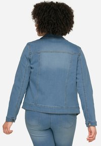 Veste en jean bleu clair à coupe classique, avec des poignets à boutons et des coutures dans le dos. Elle présente un ourlet droit et deux poches arrière visibles.
