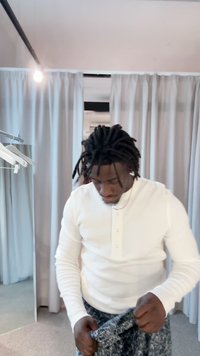 Witte henley-shirt met lange mouwen en drie knopen, gecombineerd met een broek met een patroon. Neutrale achtergrond met grijze gordijnen en een spiegel.