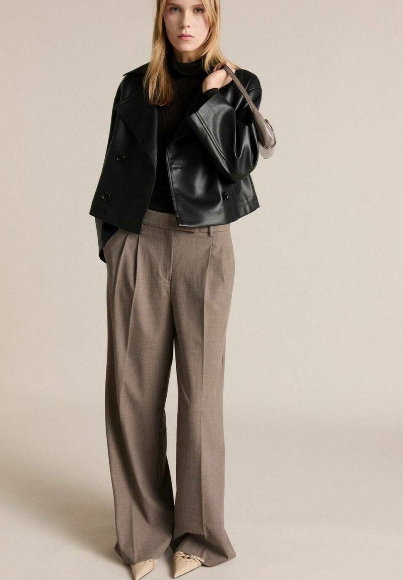 Veste en cuir noir courte, pantalons beiges longs avec plis, et escarpins nude pointus. Design simple avec une texture lisse et une coupe ajustée.