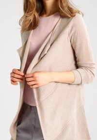 Light beige knitted cardigan with an open front, textured pattern, and long sleeves, layered over a pink top. Smooth fabric finish.