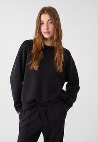 Sweatshirt noir à manches longues et col rond. Fabriqué en tissu lisse, il présente une coupe décontractée et des poignets côtelés. Il est assorti à un pantalon coordonné.