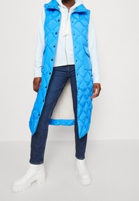 Persoon draagt een lange blauwe doorgestikte bodywarmer over een lichtblauwe hoodie, donkerblauwe spijkerbroek en dikke witte plateauboots.