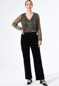 Blusa transparente de un brillante color verde oliva con mangas largas y escote en V, combinada con pantalones de pata ancha de terciopelo negro de talle alto y zapatos de tacón negros.