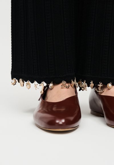 Μαύροι ριγωτοί παντελόνες με χρυσά μπιχλιμπίδια ναυτικού και θαλασσινού θέματος κατά μήκος του ποδόγυρου, φορεμένες με γυαλιστερά σκούρα καφέ δερμάτινα φλατ παπούτσια σε γυμνά πόδια.