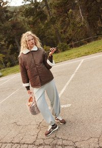 Smešani smeđi kaput s bijelim umjetnim krznom, sivi trenirka i smeđe patike. Drži ružičastu košaru s kruhom. Vanjski ambijent.