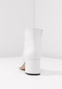 Bottine blanche à talon bloc, texture en cuir lisse, fermeture éclair au dos, et détails discrets de coutures le long des bords.