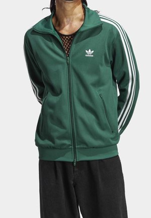 Veste zippée verte avec des rayures blanches sur les manches et le logo Adidas. Fabriquée en matériau lisse avec un col montant et des poches zippées.