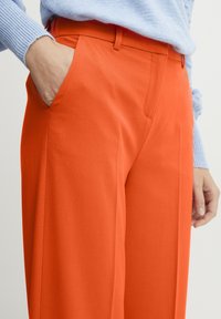Narančaste široke hlače izrađene od glatkog materijala, s bočnim džepovima i čistu waistband, u kombinaciji s svijetloružičastom pletivom puloverom.