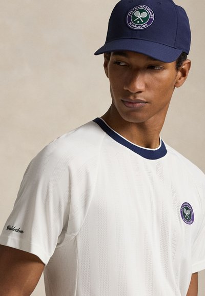 Άνδρας που φοράει λευκό μπλουζάκι Wimbledon με ναυτικό χρώμα στο τελείωμα και μπλε καπέλο μπέιζμπολ με το λογότυπο του Wimbledon, κοιτάζοντας προς τα πλάγια.