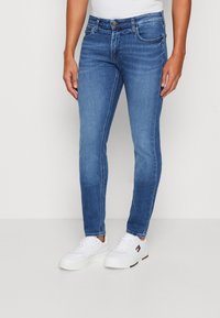 Jeans de mezclilla azul de corte ajustado con una cintura de altura media, bolsillos frontales y detalles de costura sutiles. Combinados con zapatillas blancas.
