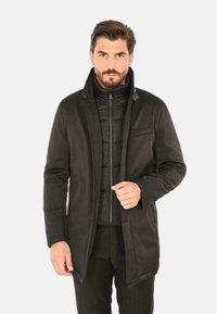 Cappotto esterno nero con colletto alto, sovrapposto a una giacca interna trapuntata. Presenta tasche frontali e accenti marroni sul colletto.