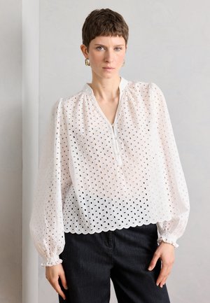 Kvinne med kort brunt hår iført gullringøreringer, en hvit bluse med hullmønster og puffermer, og svarte bukser, stående mot en ensfarget vegg.