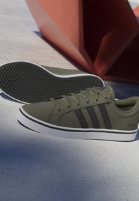 Par de zapatillas Adidas verde oliva y negras con suelas blancas colocadas sobre una superficie gris claro junto a un objeto geométrico rojo.