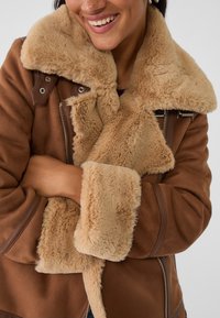 Braune Schaffelljacke mit beigem Kunstfellkragen und -manschetten. Verfügt über einen Reißverschluss und braune Akzentdetails. Weiche Textur und warmes Design.