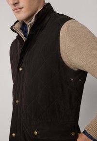 Gilet imbottito marrone con colletto alto, dotato di minuteria dorata e abbinato a un maglione beige più chiaro sotto.
