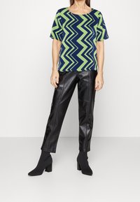 Donna che indossa una maglietta a maniche corte con motivo a zig-zag blu navy e verde, pantaloni in pelle nera e stivali alla caviglia neri con tacco blocco.