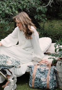 Blomstermønstret tote-bag i grønne og beige farver, med brune stropper, holdbart stof, hvilende på en beige pude nær en klapvogn i en naturlig omgivelser.