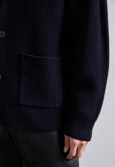 Tumedast sinine paksu kudumisega kardigan, millel on ees üle õla tasku ja mustad nööbid, kantud mustade pükste peal, näidates alakeha ja paremat kätt.