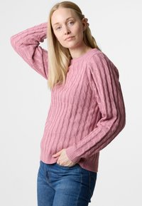 Rosa Kabelstrickpullover mit geripptem Saum und Ärmeln, der ein strukturiertes Design aufweist. Getragen mit blauen Jeans, die Hände auf den Hüften.
