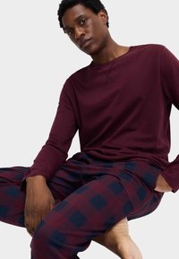 Homme assis en tailleur portant un haut à manches longues bordeaux et un pantalon à carreaux bleu marine et bordeaux sur un fond blanc uni.