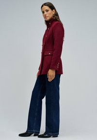 Chaqueta entallada burdeos con un cuello alto, botones dorados, bolsillos de solapa, combinada con jeans de mezclilla oscura de pierna ancha y botas de tacón negras.