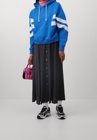 Persoon in een blauwe hoodie met witte strepen, een zwarte knopenrok, witte sokken, zwarte sneakers en met een glanzende roze handtas.