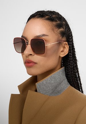 Donna con capelli intrecciati che indossa occhiali da sole quadrati oversize e un cappotto colore cuoio con colletto grigio, rivolta leggermente a sinistra.