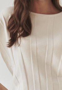 Top beige chiaro realizzato in tessuto morbido, con pieghe verticali e scollatura ampia, dalla consistenza liscia e vestibilità comoda.