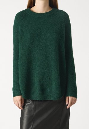 Personne portant un pull à manches longues vert foncé duveteux et une jupe en cuir noire, debout devant un fond blanc uni.