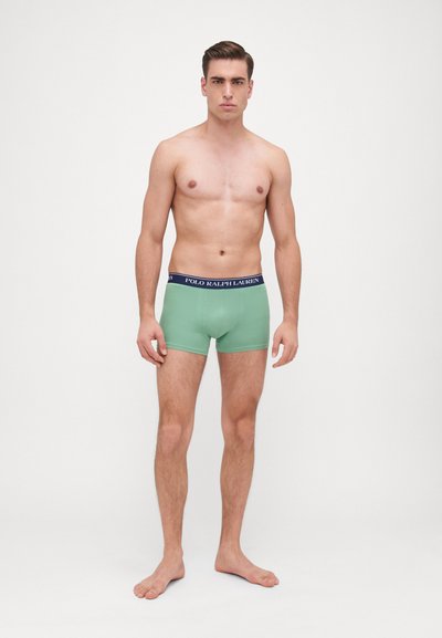 Zöld pamut boxer nadrág sötétkék derékrésszel, amelyen a "POLO RALPH LAUREN" logó látható. A modell mezítláb áll egy semleges háttér előtt.
