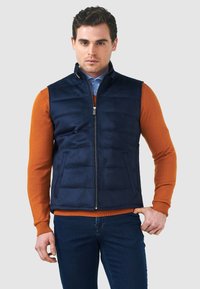Marineblau gesteppte Weste mit hohem Kragen, Frontreißverschluss und zwei Seitentaschen, getragen über einem orangefarbenen Pullover und blauen Jeans. Glatte Textur.