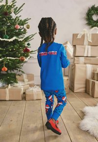 Bambino con capelli intrecciati che indossa un pigiama di Spider-Man tiene un regalo vicino all'albero di Natale decorato e ai pacchi regalo sul pavimento di legno.