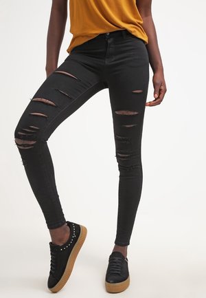 Personne portant un jean skinny noir déchiré, un haut jaune moutarde et des baskets noires avec des semelles marron, debout devant un fond uni.