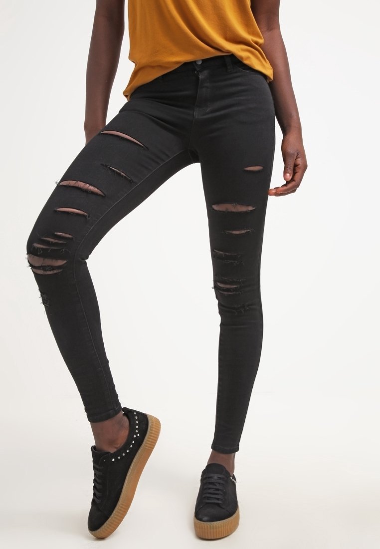 Personne portant un jean skinny noir déchiré, un haut jaune moutarde et des baskets noires avec des semelles marron, debout devant un fond uni.