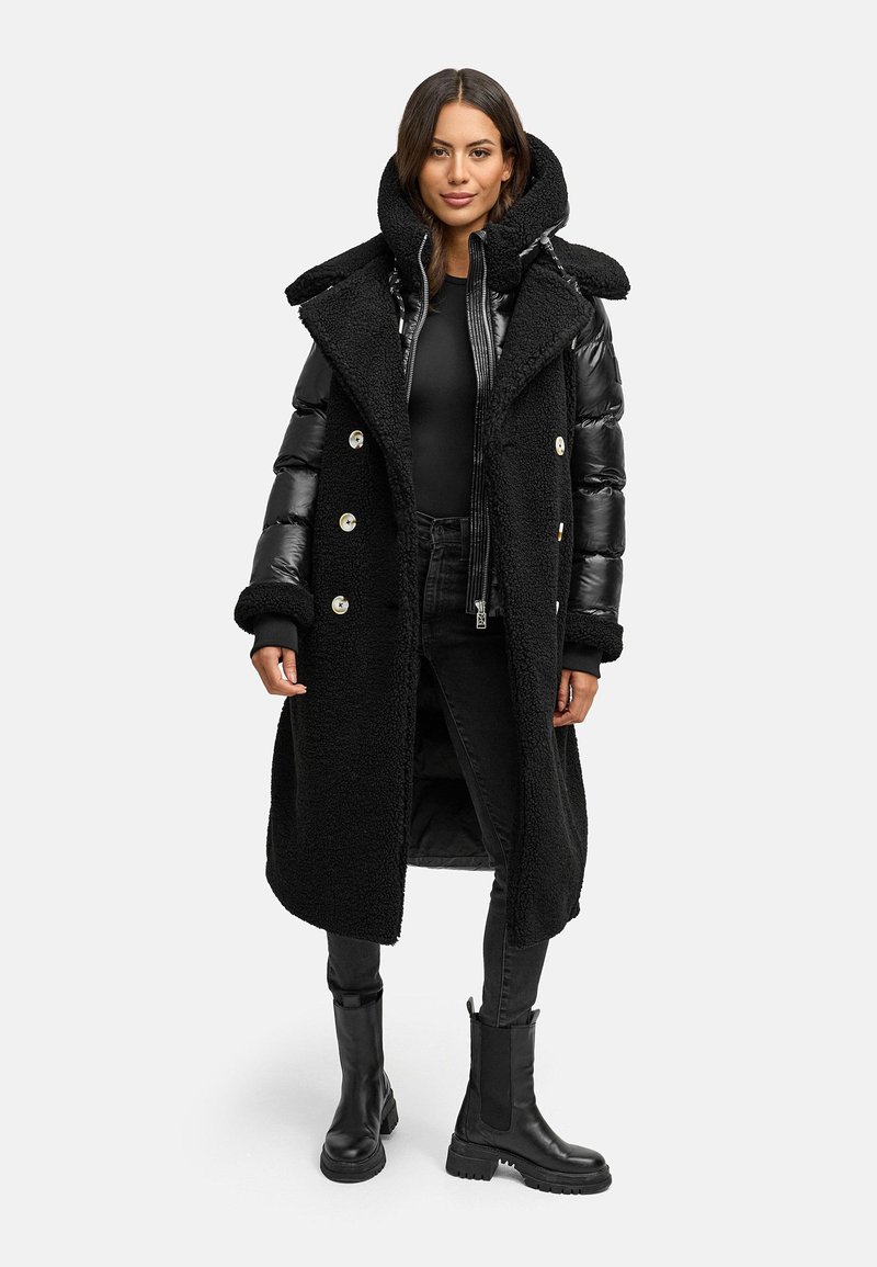 Cappotto lungo nero con un esterno morbido e texture, maniche gonfie lucide, bottoni doppi, e un ampio cappuccio, abbinato a jeans neri e stivali.