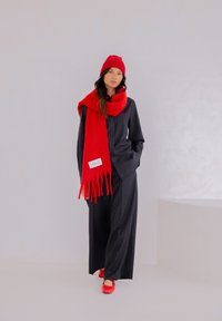 Model in een zwart gestreept pak met wijde pijpen, rode gebreide muts en bijpassende oversized sjaal met franjes, en rode platte schoenen.