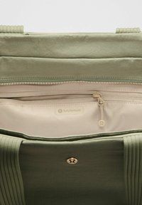 Zöld szövet táska redős textúrával, arany cipzárral, bézs bélésbéll és egy kis Lululemon logóval az elején.