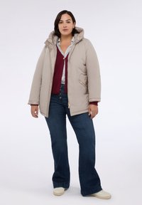 Písková prošívaná bunda s kapucí, zlatými doplňky, předními kapsami, nošena přes vínový svetr a světlemodrou košili, v kombinaci s tmavými džínami.