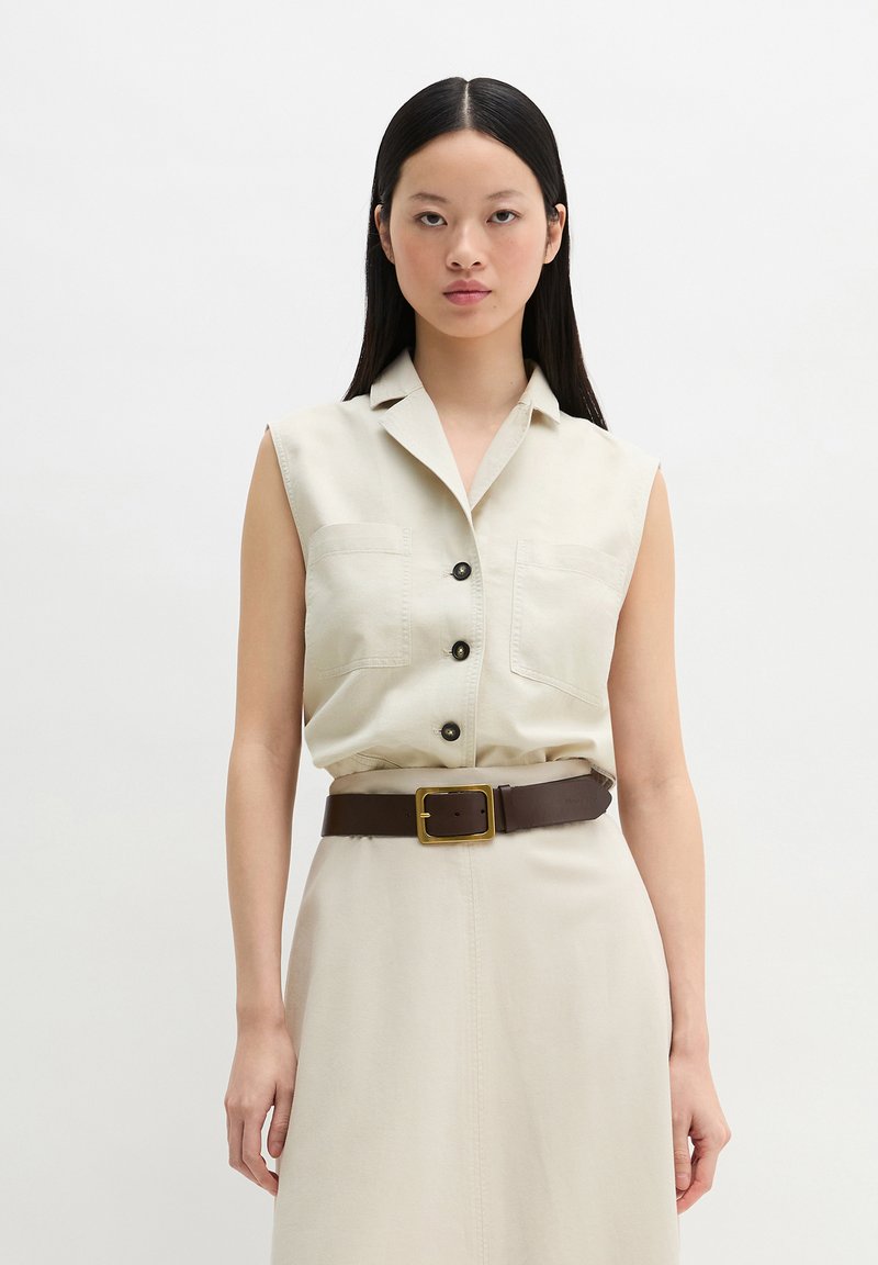 Femme aux longs cheveux noirs portant une robe beige sans manches boutonnée avec poches plaquées et ceinture marron sur un fond clair uni.