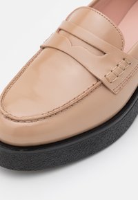 Mocassin en cuir beige avec détails cousus et semelle texturée noire sur fond blanc, gros plan sur la partie avant de la chaussure.