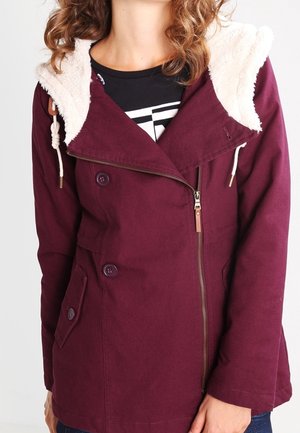 Femme portant une veste bordeaux avec capuche doublée en peau lainée, fermeture éclair diagonale, poche à rabat boutonnée et détails à boutons sur un haut noir.